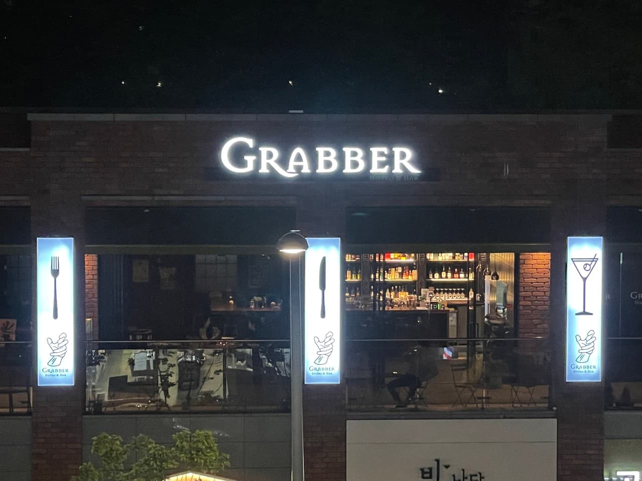 야간의 벽돌 외벽 위로 포크와 칵테일 아이콘 조명이 켜져 있고 창문 너머로 술병이 진열된 선반과 길게 늘어선 바 카운터 좌석이 보임