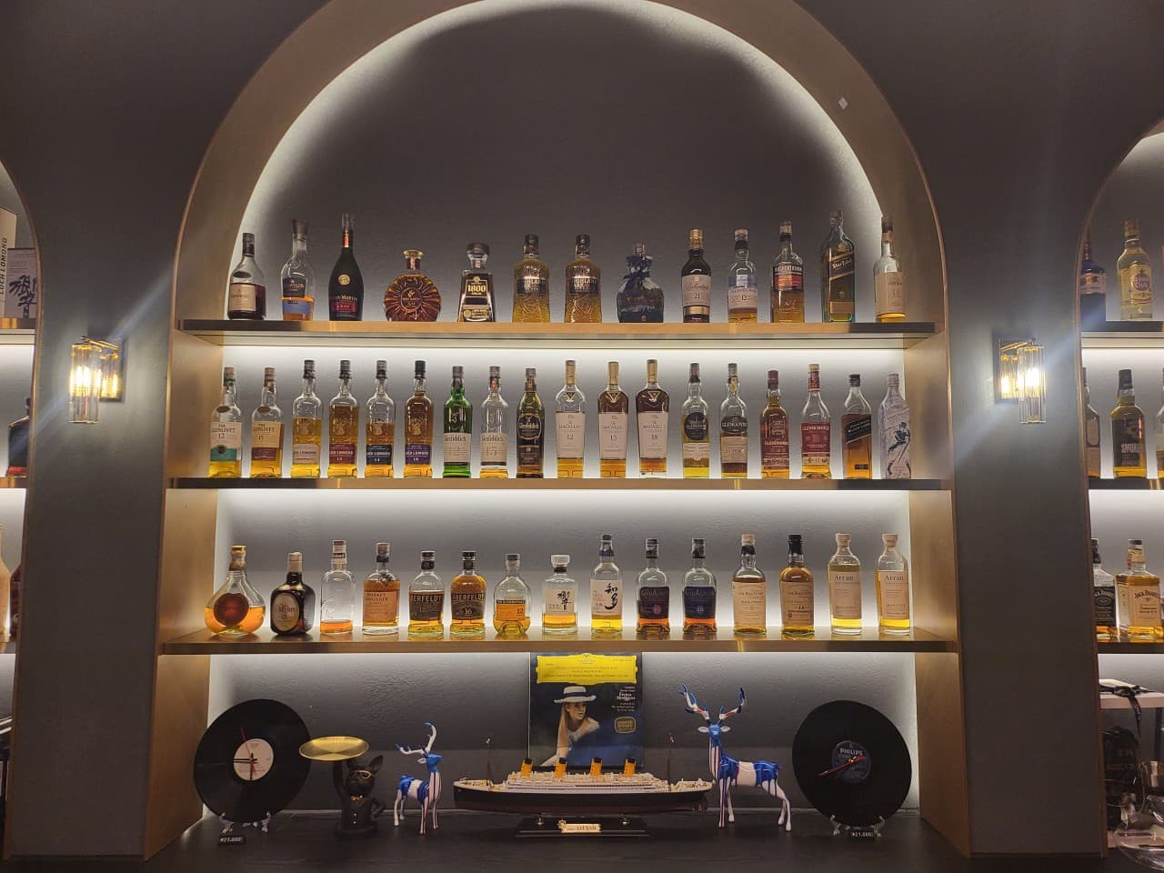 아치형 벽면 선반의 간접 조명 아래 정렬된 위스키 병들과 하단에 배치된 LP 레코드 및 모형