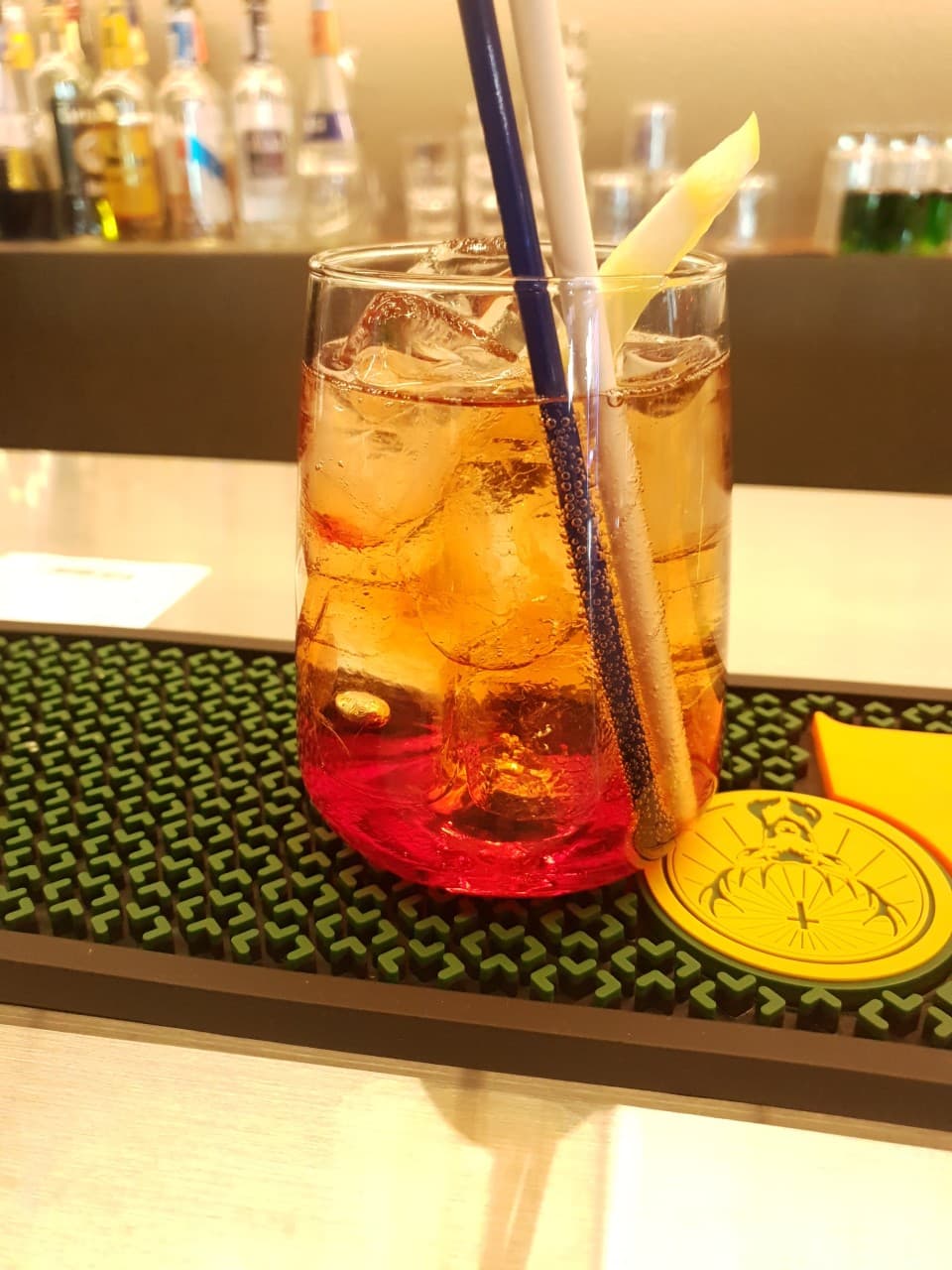 술병이 진열된 바 카운터 위 초록색 고무 매트에 놓인 얼음과 레몬 조각이 담긴 하이볼 잔