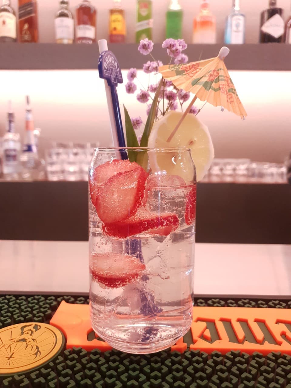 슬라이스 딸기와 레몬, 종이 우산이 꽂힌 투명한 하이볼 잔이 놓인 바 카운터와 배경에 진열된 술병들