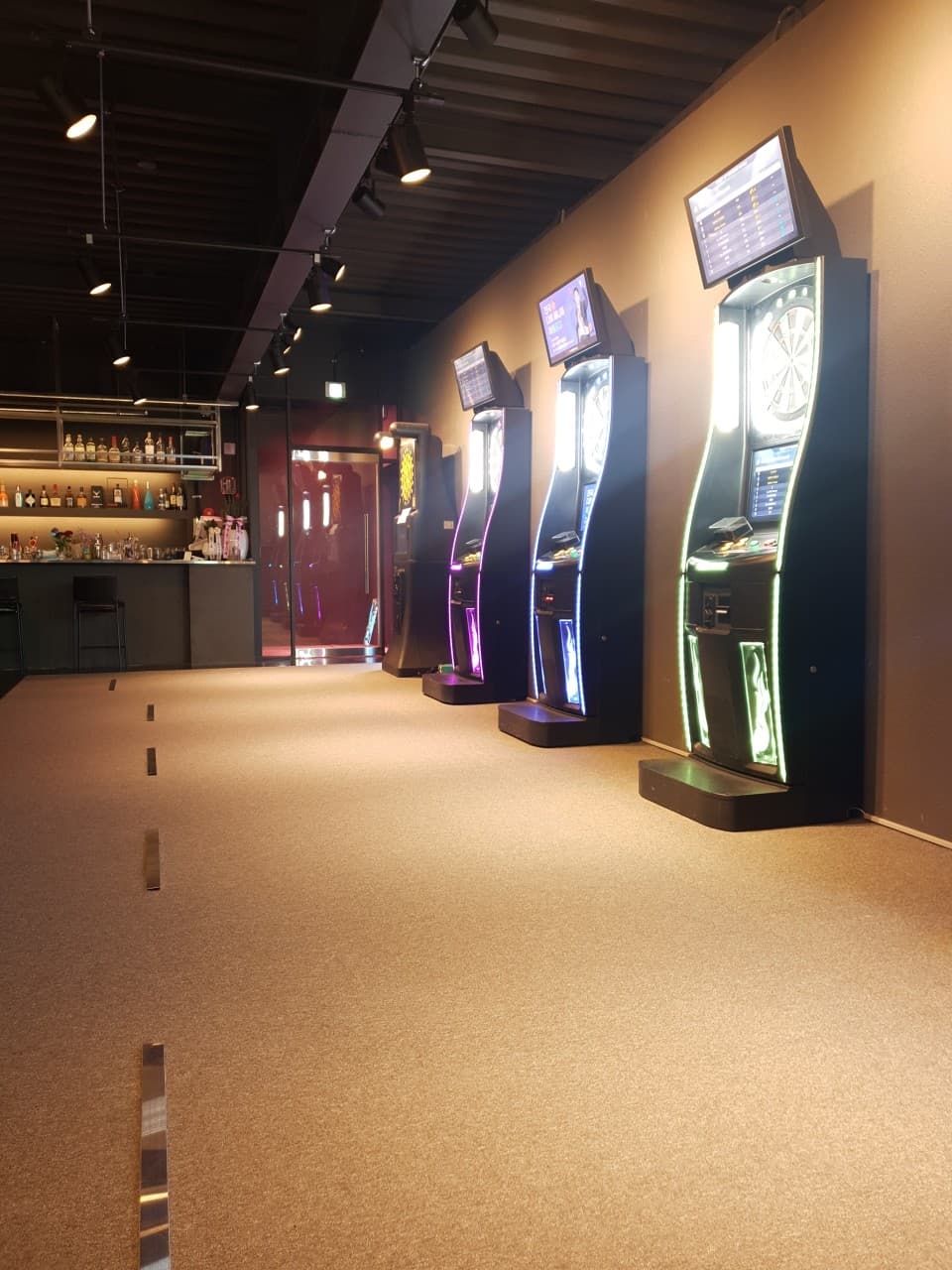 어두운 실내 스포트라이트 아래 나란히 배치된 다트 머신들과 술병이 진열된 바 카운터 및 좌석