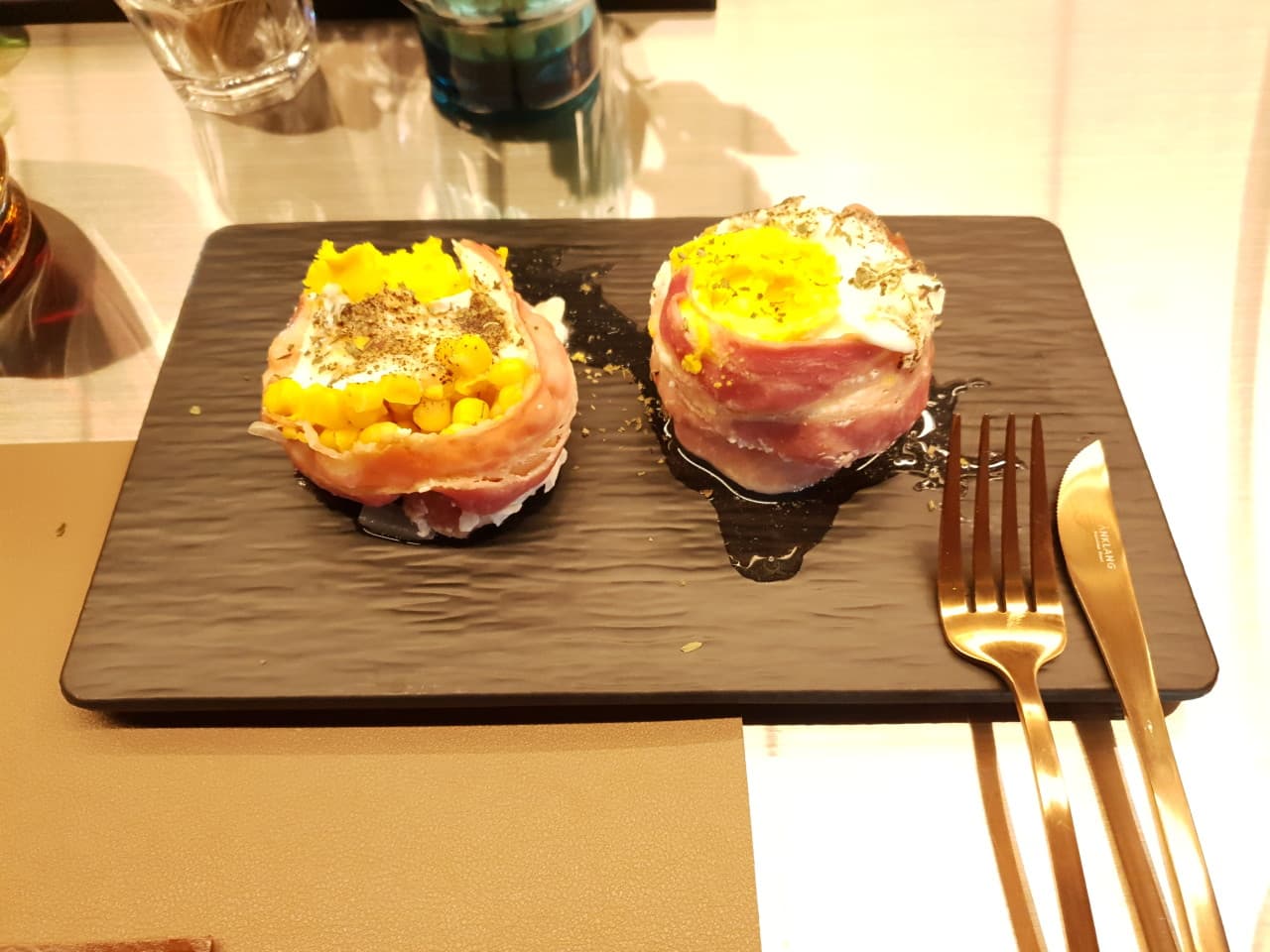 검은색 사각 플레이트 위 베이컨으로 감싼 달걀 요리 두 점과 금색 포크, 나이프가 놓인 1인석