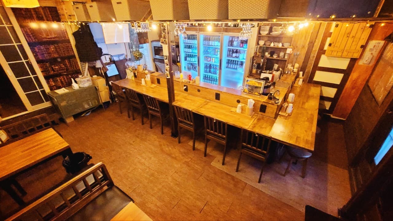 황색 조명이 켜진 긴 목재 카운터와 1인용 의자들이 일렬로 배치된 바 테이블. 뒤편에는 주류 냉장고와 식기가 수납된 선반이 위치함.