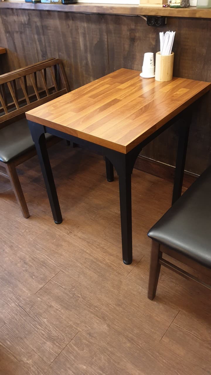 나무 결이 보이는 사각형 테이블과 검정 가죽 의자. 벽면 쪽에는 종이컵과 나무 젓가락이 담긴 통이 비치됨.