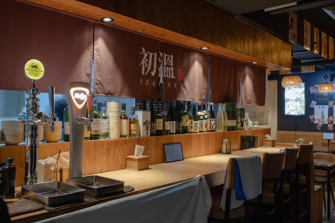 갈색 가림막 아래 긴 목재 카운터와 높은 의자가 일렬로 배치된 공간, 선반 위에 진열된 여러 술병과 생맥주 탭