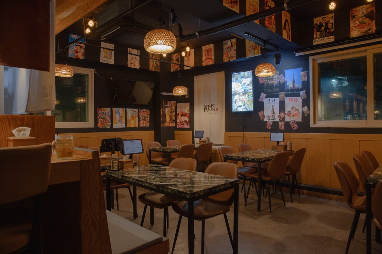 주황빛 조명이 비추는 목재 카운터석과 대리석 무늬 테이블. 벽면의 일본식 포스터들과 좌석마다 비치된 태블릿 형태의 주문 기기.