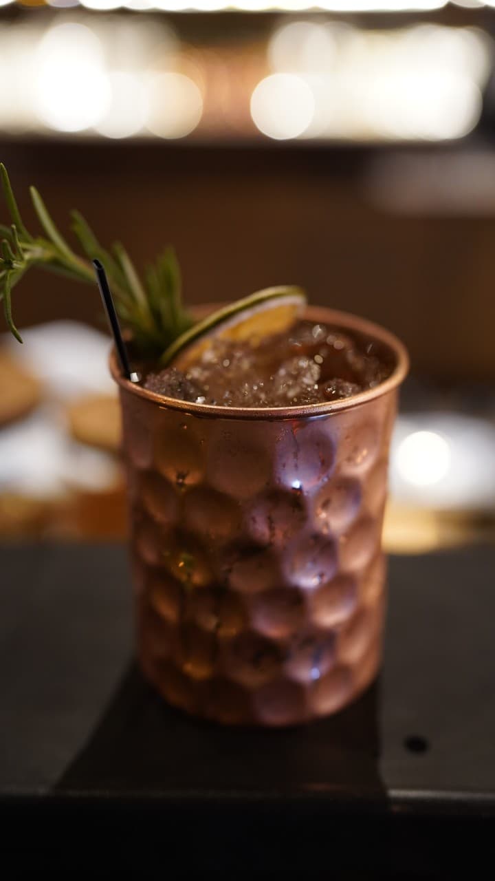 어두운 조명 아래 바 테이블 위 로즈마리와 말린 라임이 꽂힌 엠보싱 구리 잔 속 얼음 가득한 음료