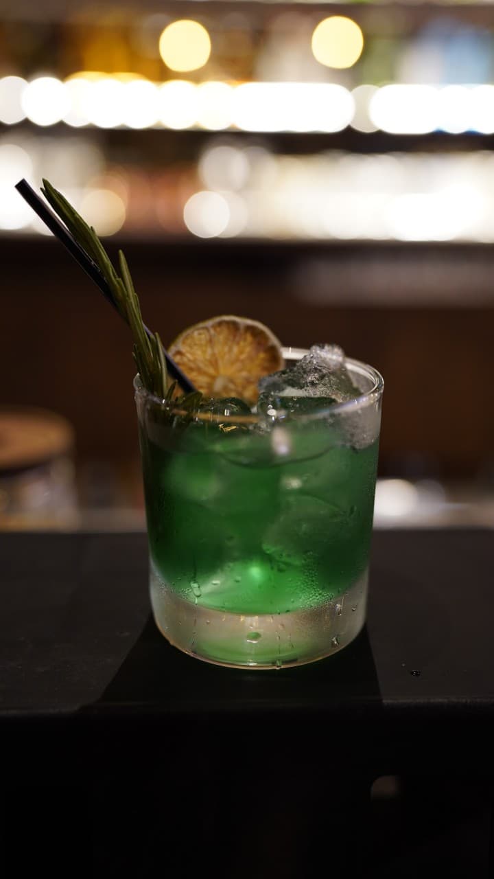 어두운 조명의 바 카운터 위 얼음 가득한 초록색 칵테일과 말린 라임, 로즈마리 허브 장식