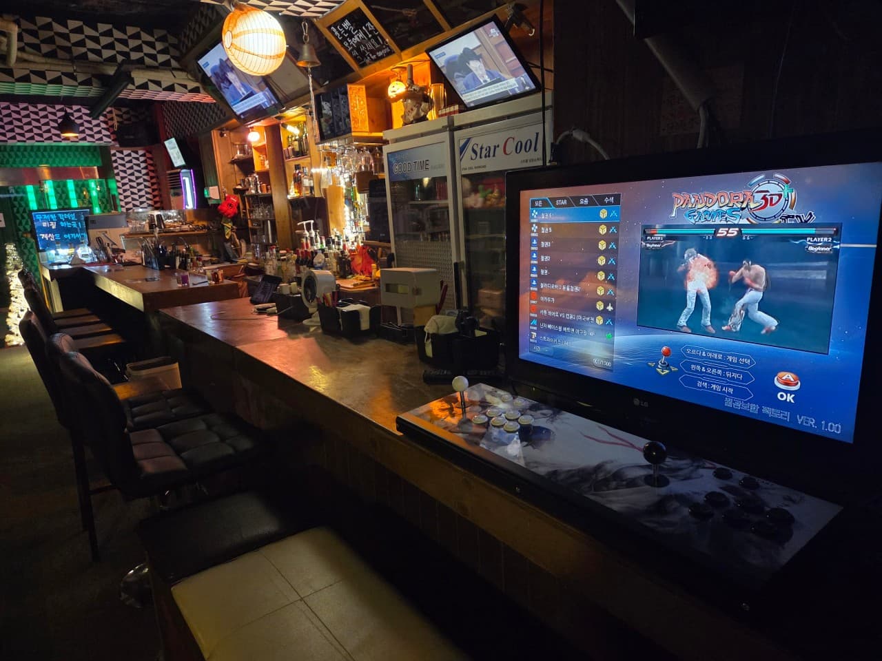 어두운 조명 아래 아케이드 게임기가 설치된 나무 카운터와 일렬로 배치된 1인용 의자, 술병이 진열된 선반