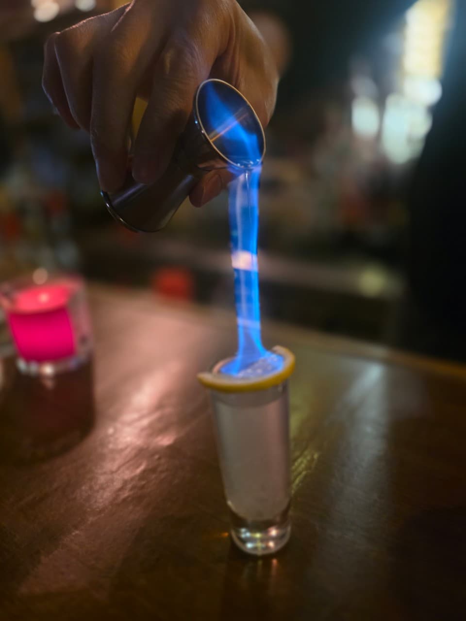 어두운 목재 카운터 위 분홍색 촛불 옆 지거에서 샷잔으로 쏟아지는 푸른 불꽃과 레몬 조각이 얹어진 술