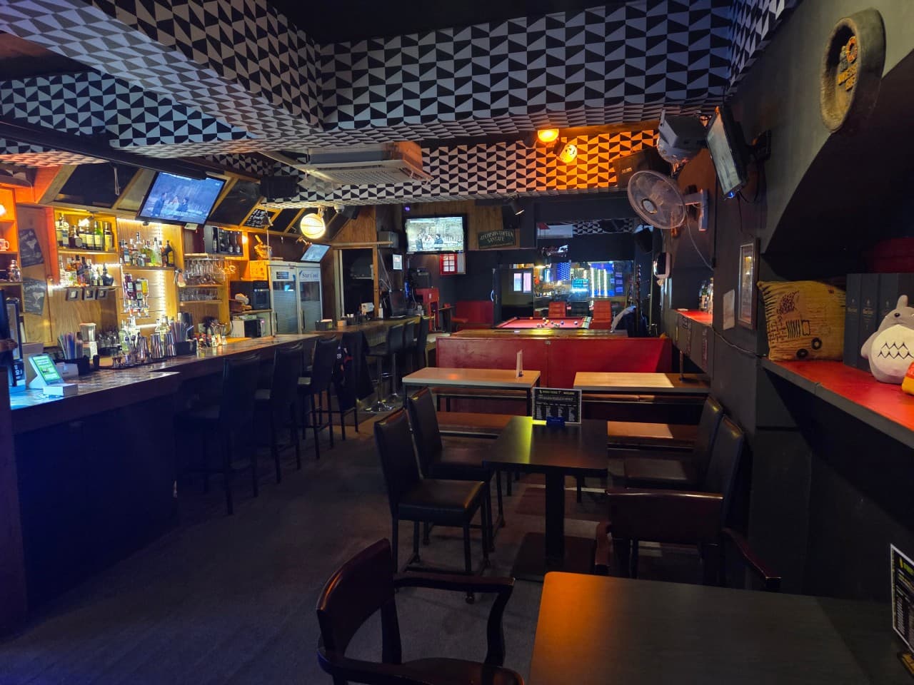기하학적 패턴의 천장 아래 긴 바 카운터와 높은 의자, 안쪽에 배치된 당구대와 어두운 조명의 1인용 테이블 좌석
