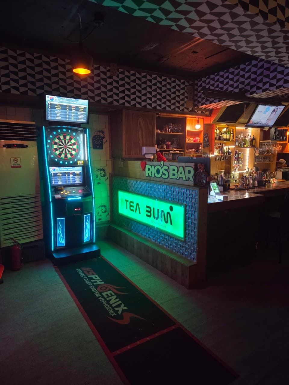 푸른 조명의 전자 다트 기계와 다양한 술병이 진열된 일자형 나무 바 테이블 및 카운터석