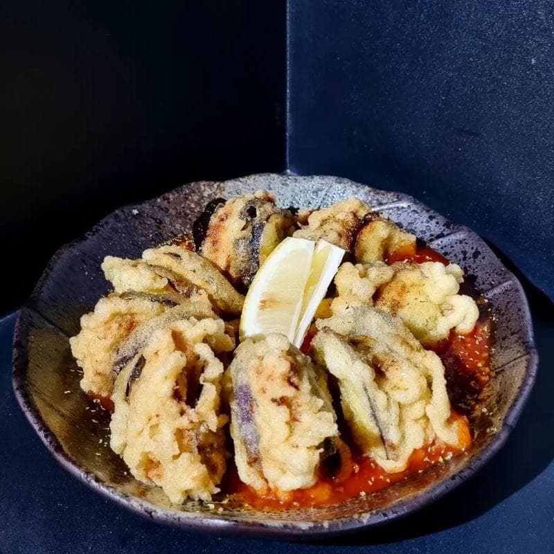 어두운 무채색 배경 속 붉은 소스가 깔린 접시 위에 나란히 놓인 가지 튀김과 단면을 보인 레몬