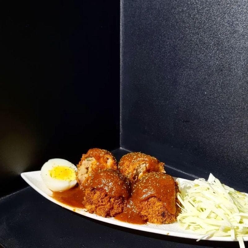 어두운 배경 속 흰 접시에 담긴 갈색 소스를 곁들인 튀김 요리와 반으로 자른 삶은 계란, 채 썬 양배추