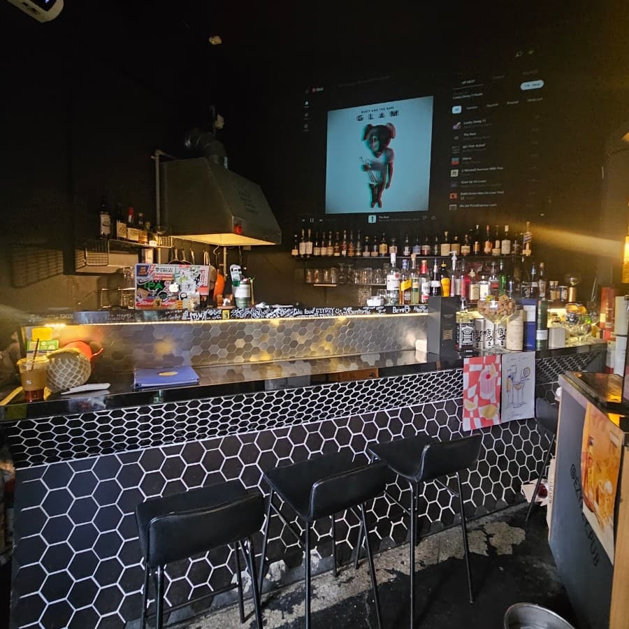 어두운 조명 아래 육각 타일 바 테이블과 높은 의자가 배치된 공간, 벽면에 진열된 다양한 술병들과 영상이 나오는 대형 스크린