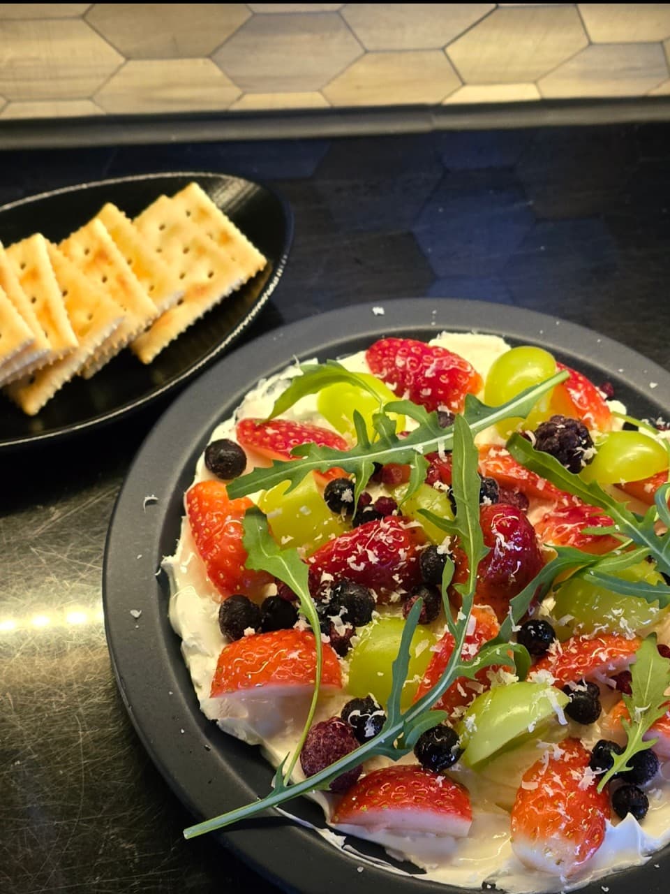 어두운 질감의 카운터 위 딸기, 청포도, 루꼴라가 곁들여진 크림치즈 플레이트와 소량의 크래커 한 접시