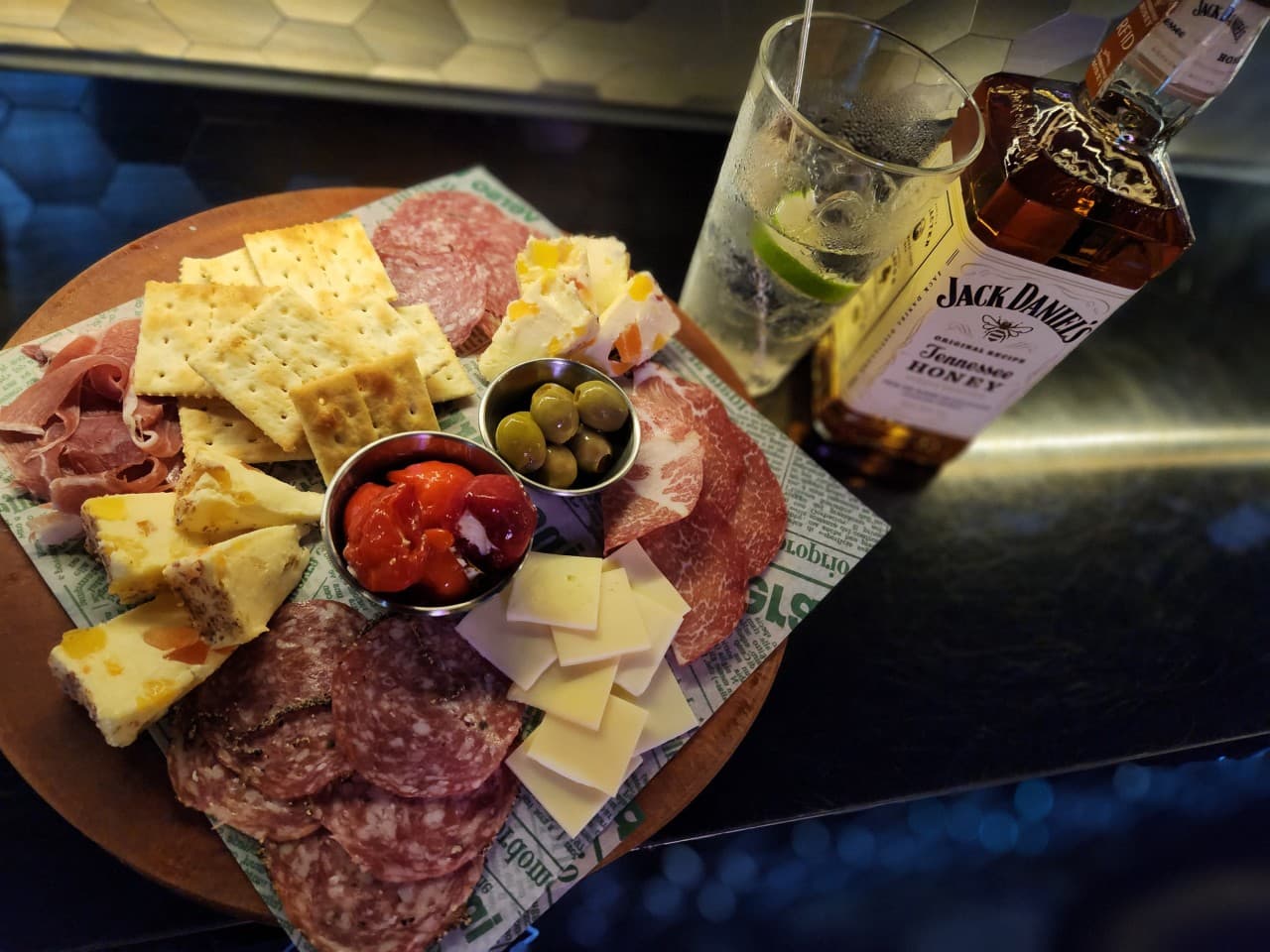 어두운 조명 아래 바 테이블 위 크래커와 치즈, 살라미가 담긴 플래터와 위스키 병, 라임을 넣은 하이볼 잔