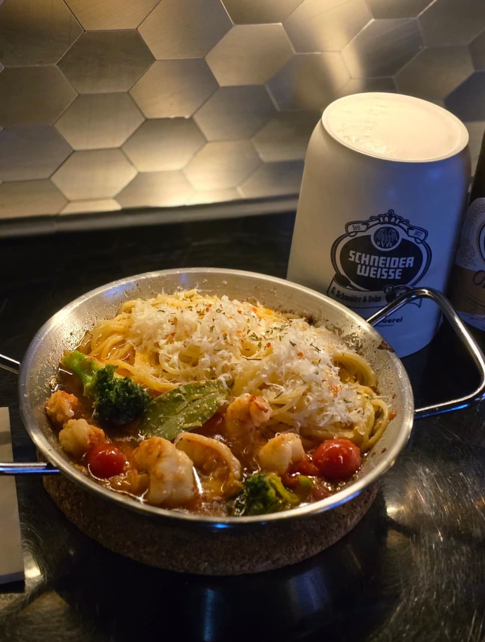 육각형 타일 벽 앞 검은 테이블 위 치즈를 올린 새우 파스타와 거품이 담긴 도자기 맥주잔
