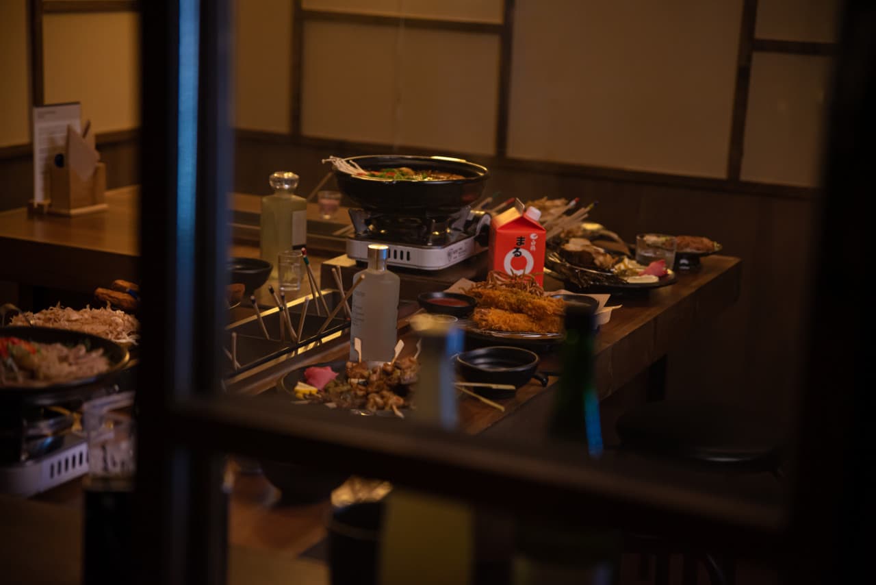 어두운 조명 아래 목재 카운터 위 전골 냄비와 붉은색 사케 팩, 꼬치 안주와 술병이 차려진 1인석 바 테이블