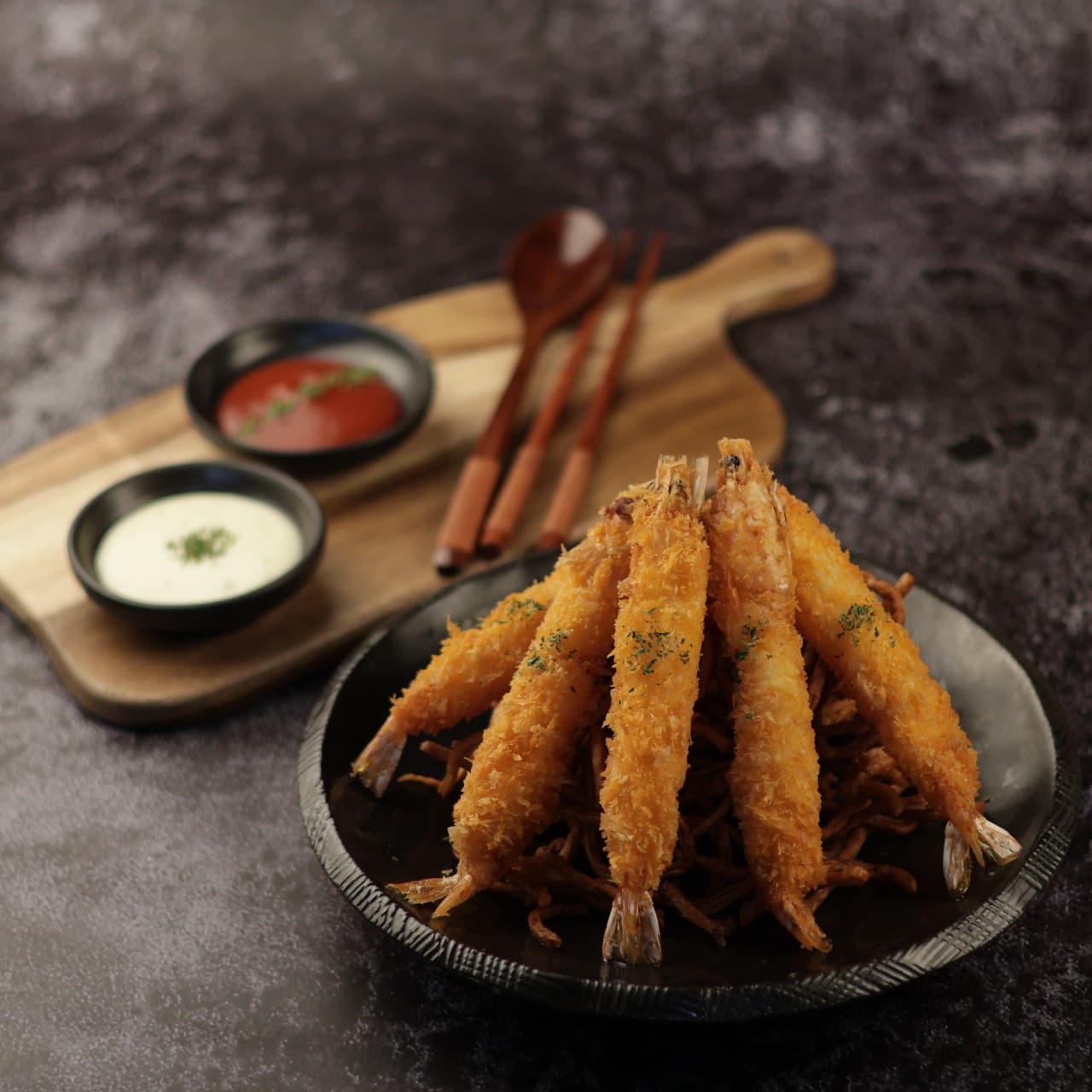 어두운 배경 속 검은 접시에 담긴 새우튀김과 나무 도마 위 두 종류 소스, 목제 수저가 놓인 1인 상차림