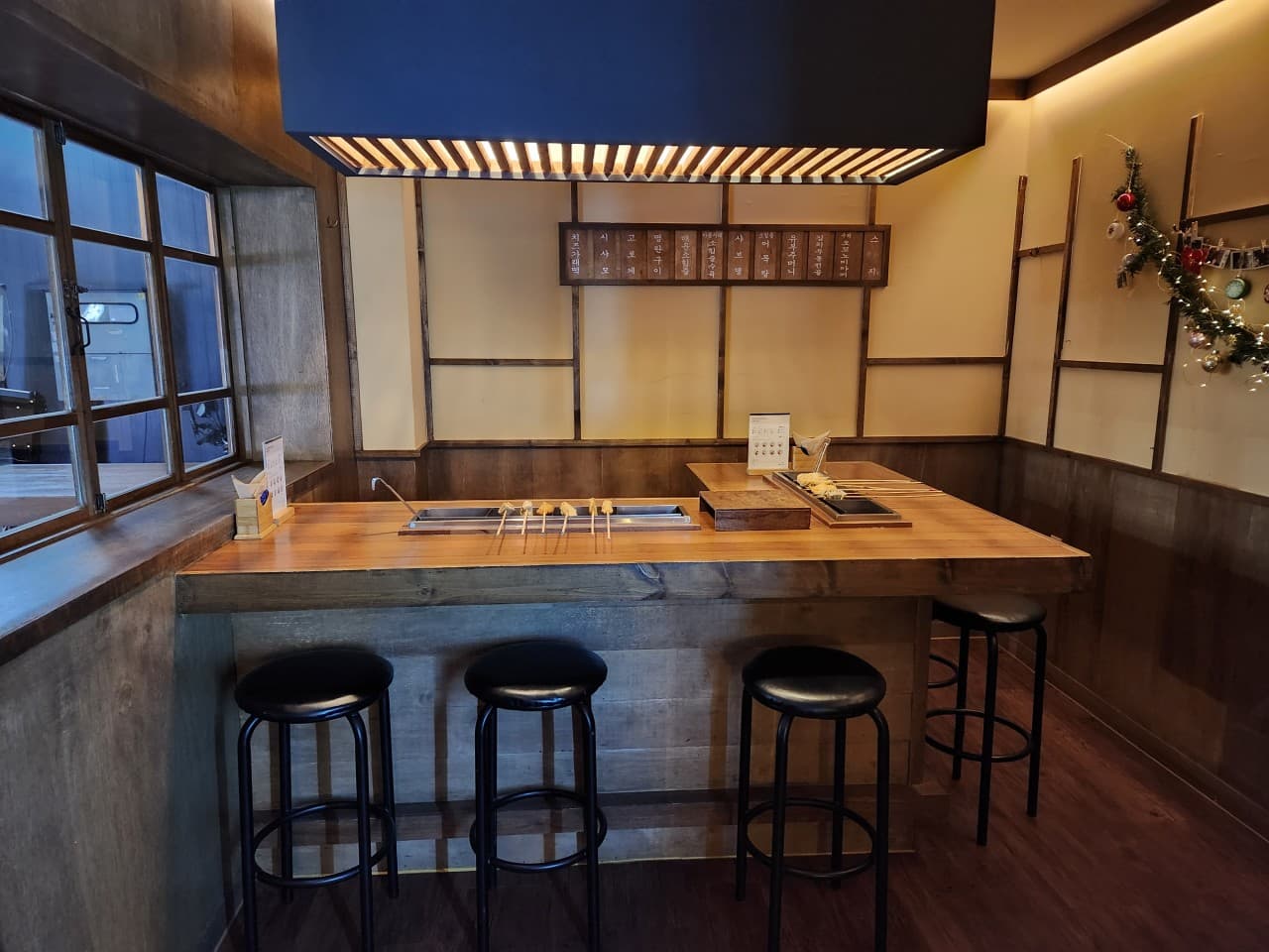 목재 카운터 테이블 위 어묵 조리기와 꼬치, 그 앞에 일렬로 배치된 1인용 검은색 바 스툴과 상단 사각형 조명