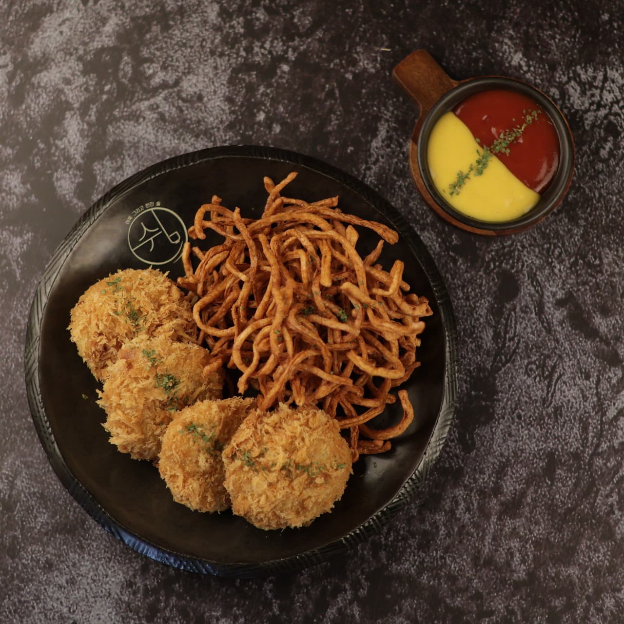 어두운 배경 위 검은 접시에 담긴 네 개의 둥근 튀김과 가느다란 어묵 채 튀김, 두 가지 소스가 담긴 작은 종지