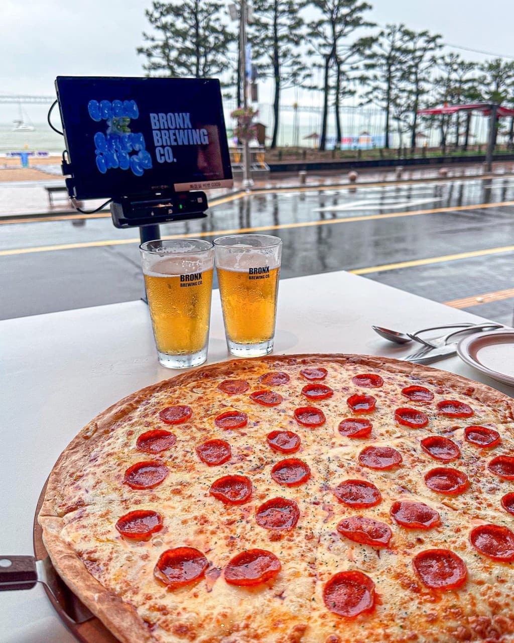 해변 도로와 바다가 보이는 야외 테이블 위 놓인 대형 페퍼로니 피자와 태블릿 메뉴판 옆 생맥주 두 잔