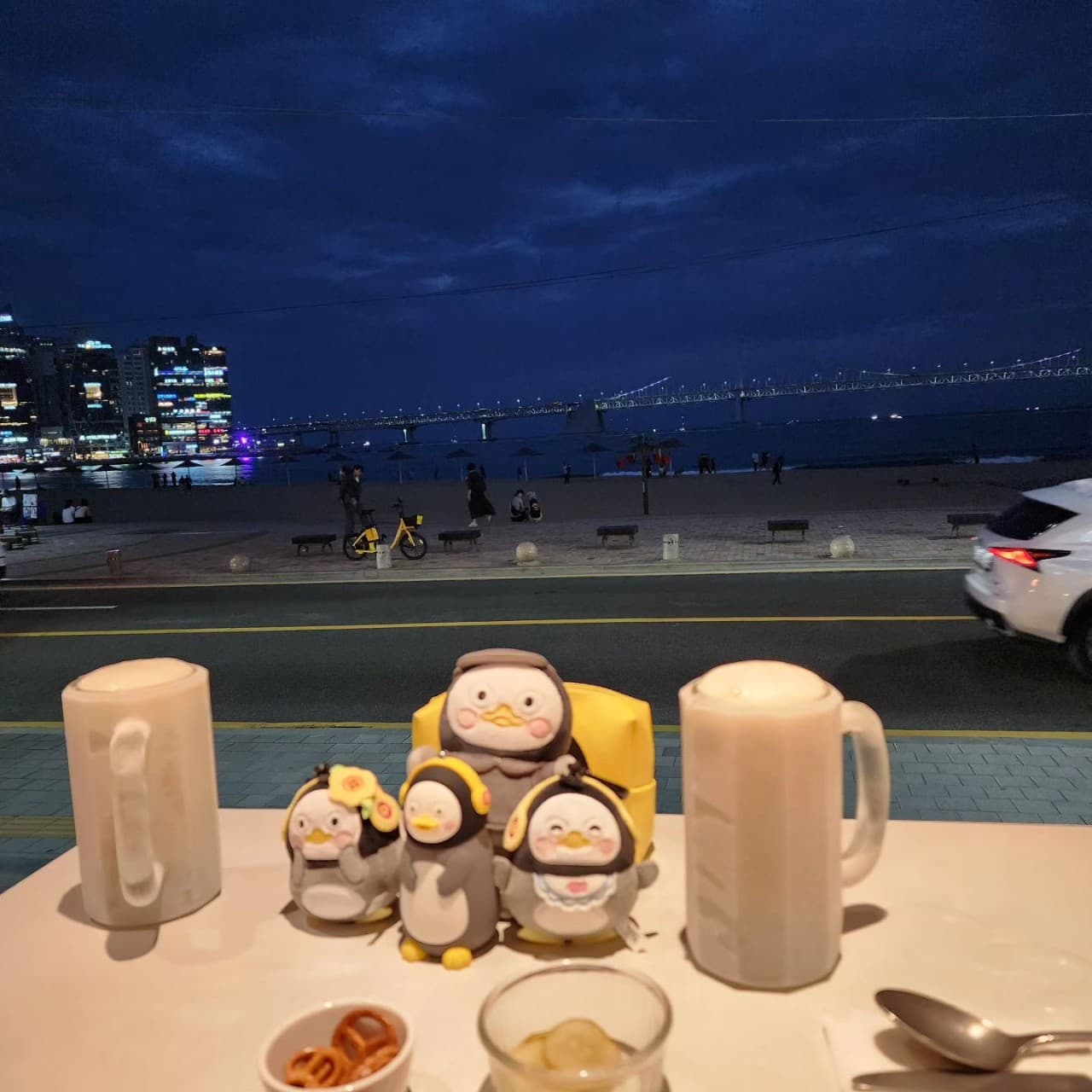 어두운 밤바다와 대교 야경이 내다보이는 창가 자리에 놓인 거품 올라온 맥주 두 잔과 펭귄 캐릭터 소품들