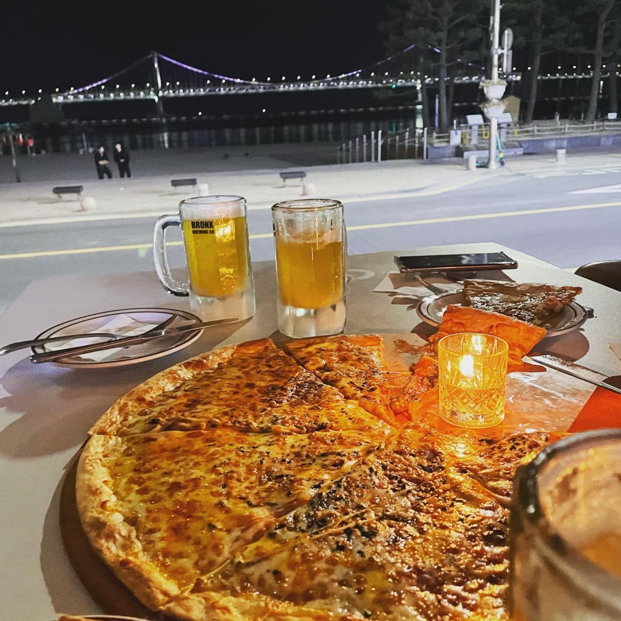 보라색 조명이 켜진 대교와 밤바다를 마주한 테이블 위 대형 피자와 맥주잔 그리고 촛불