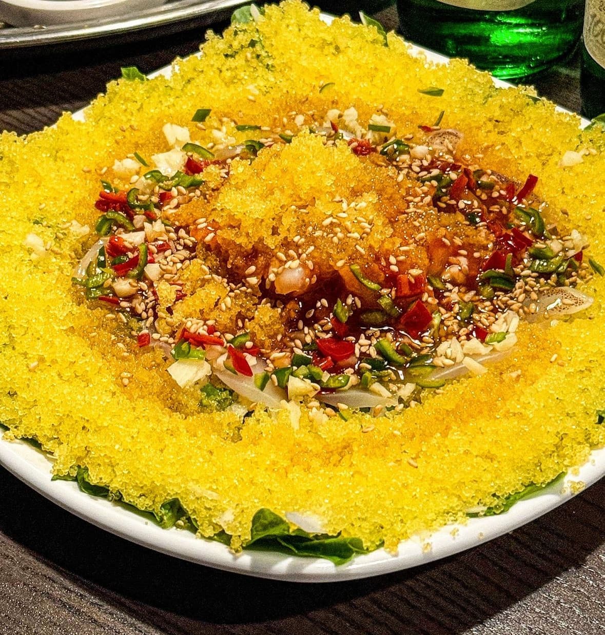 흰 접시 위 노란 날치알과 잘게 썬 고추, 참깨가 뿌려진 요리와 어두운 목재 테이블 위 초록색 술병