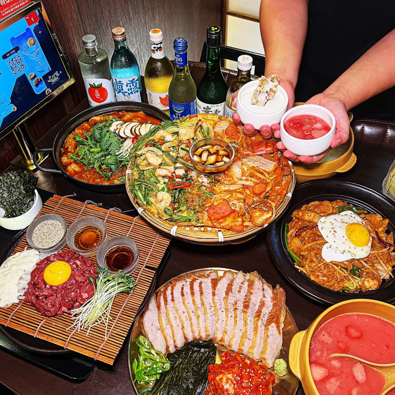 노른자를 얹은 육회와 수육, 전 등 다양한 안주가 차려진 테이블 위 나란히 놓인 여러 술병과 작은 그릇을 든 손