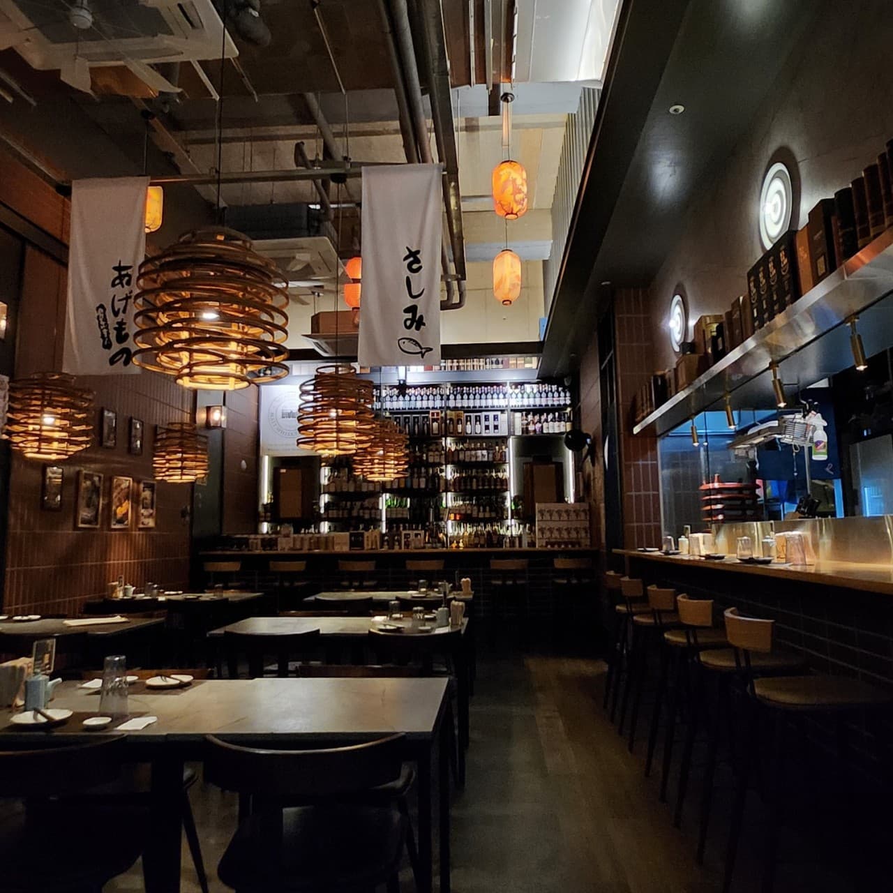 주황색 조명 아래 놓인 긴 목재 바 카운터와 1인용 높은 의자들. 벽면을 가득 채운 술병 진열장과 천장의 일본어 현수막