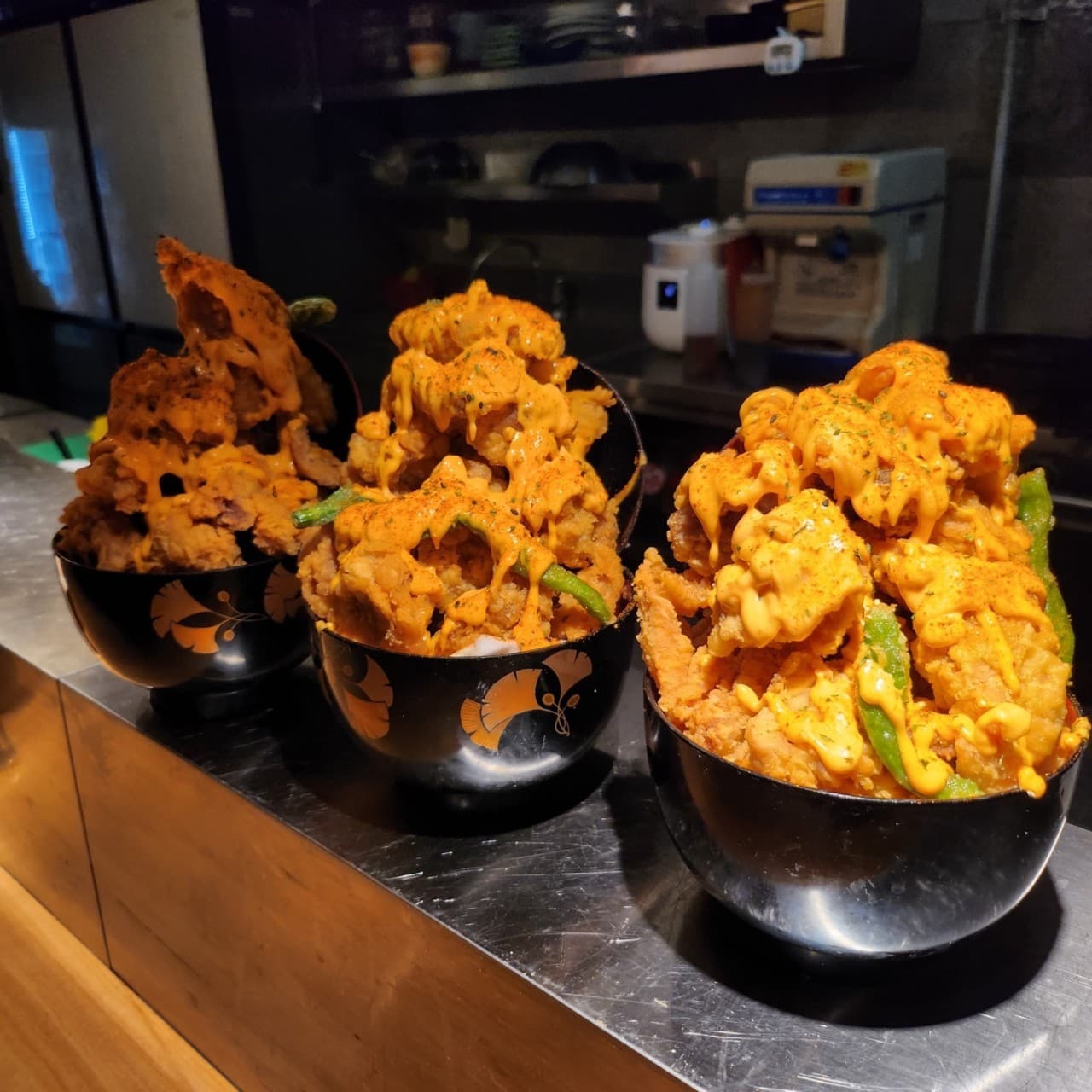 어두운 조명의 스테인리스 바 테이블 위 주황색 소스가 뿌려진 세 그릇의 높게 쌓인 튀김과 꽈리고추