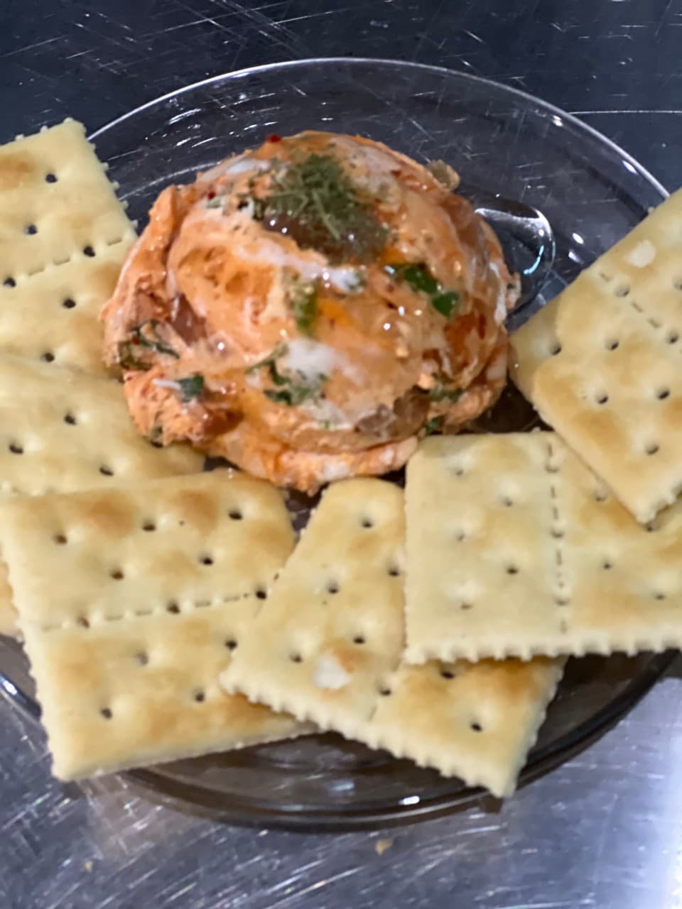 투명한 유리 접시 위 허브 가루를 얹은 주황색 스프레드 한 덩이와 그 주변을 둘러싼 여러 장의 사각형 크래커