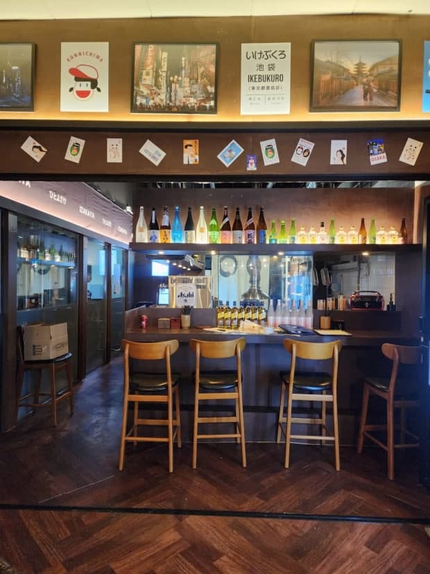 일렬로 늘어선 술병 아래 네 개의 목재 의자가 배치된 바 형태의 카운터 좌석. 벽면에는 일본풍 포스터가 부착된 어두운 조명의 실내