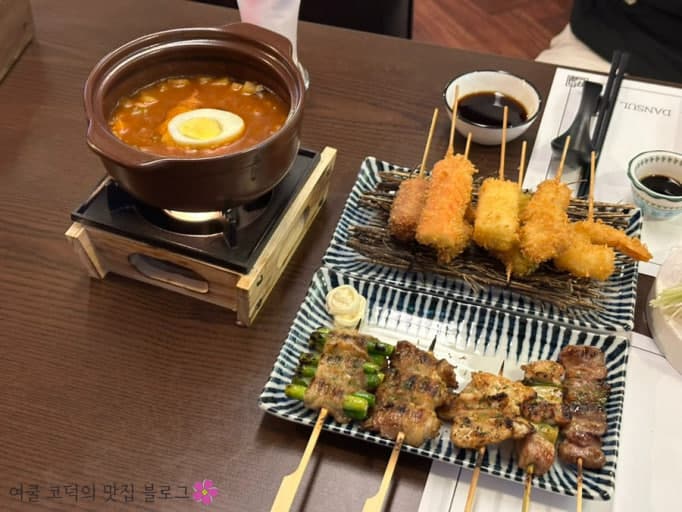 1인용 화로 위 뚝배기 국물 요리와 두 개의 접시에 담긴 고기 꼬치구이, 튀김 꼬치 및 간장 종지가 놓인 목재 테이블