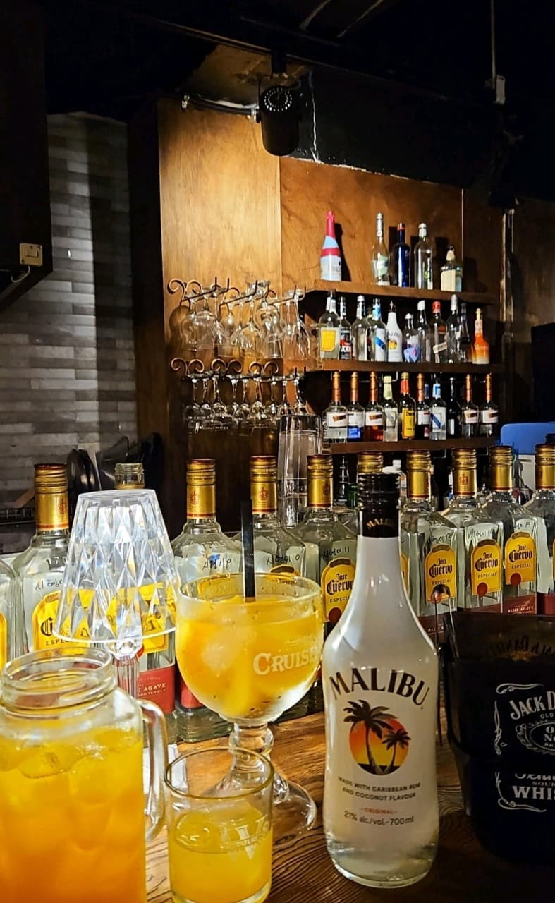 어두운 조명 아래 목재 바 테이블 위 말리부 병과 노란색 칵테일, 벽면 선반에 진열된 다양한 주류와 글라스들