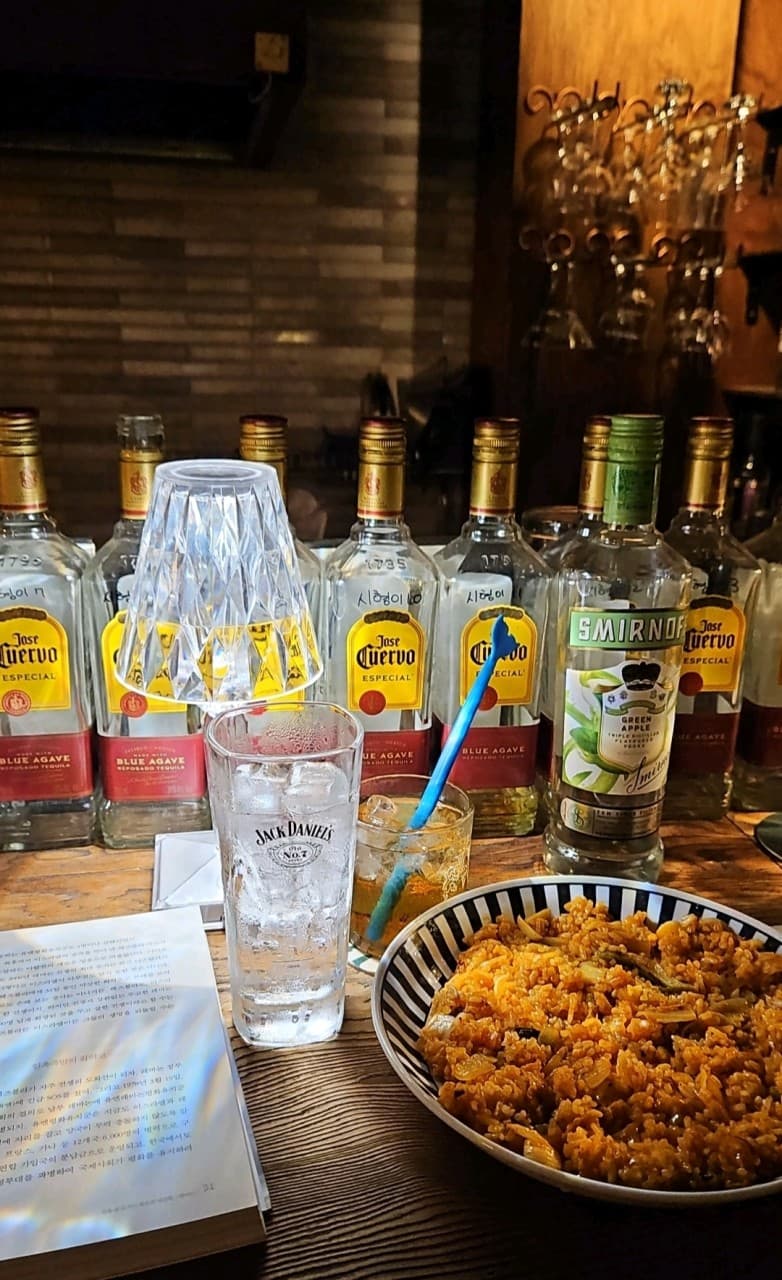 조명이 켜진 나무 카운터 위 펼쳐진 책과 볶음밥, 얼음이 담긴 잔 옆으로 나열된 여러 병의 테킬라와 보드카