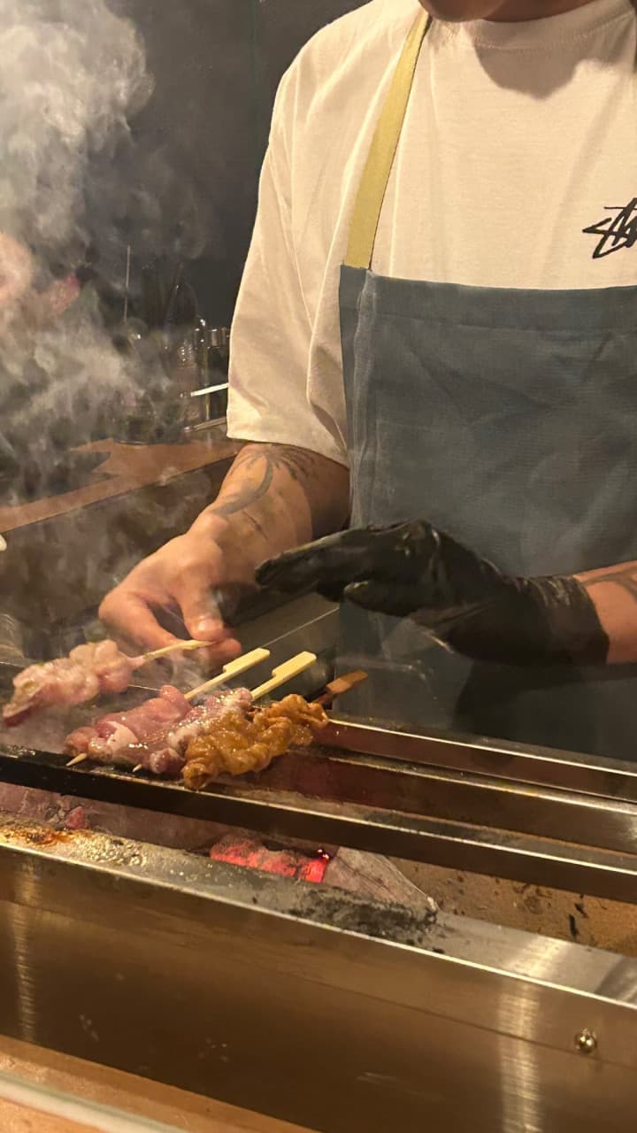 숯불 위에서 연기를 내며 익어가는 닭꼬치와 이를 조리하는 요리사의 손길, 카운터 좌석 정면의 모습