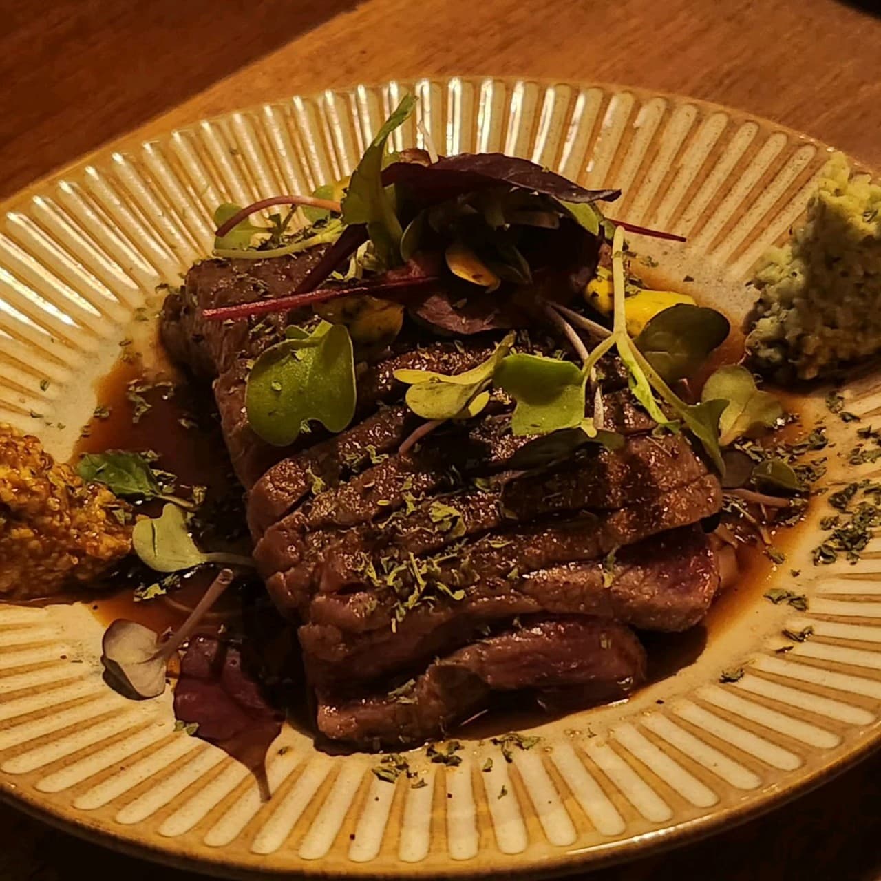 어두운 조명 아래 나무 테이블 위 소고기 구이와 채소 고명, 와사비가 곁들여진 1인용 접시