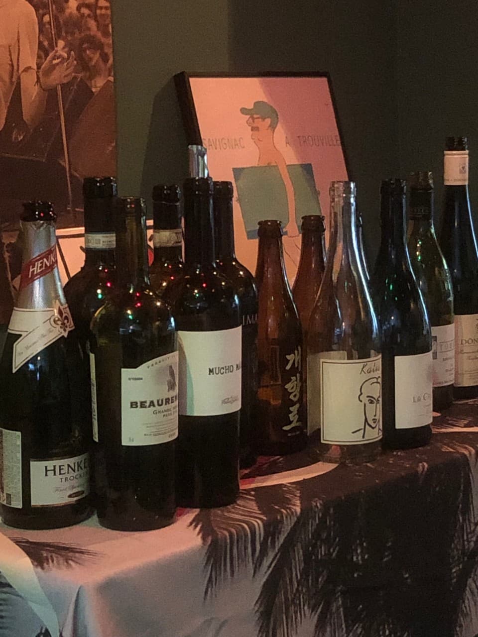 어두운 조명 아래 야자수 무늬 식탁보 위로 나열된 다양한 종류의 와인병과 맥주병, 벽면에 세워진 인물 포스터