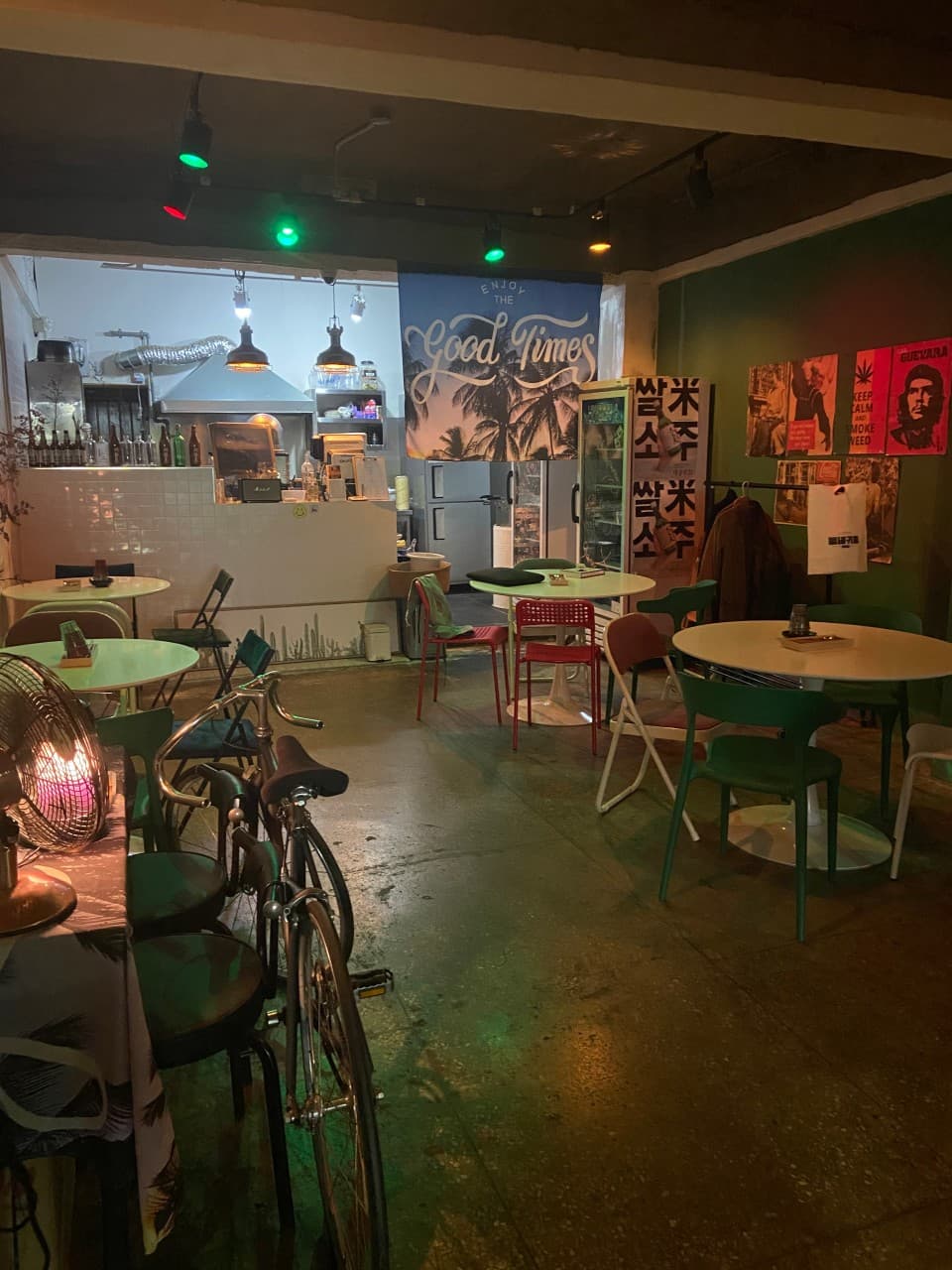 녹색 벽면 포스터 아래 소형 원형 테이블과 1인용 의자가 놓인 어두운 실내와 천장의 유색 조명