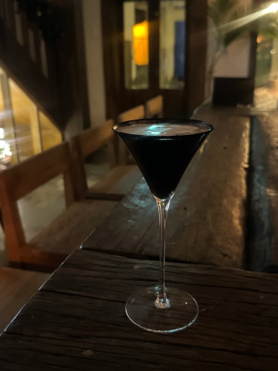 거친 질감의 나무 카운터 위 검은색 칵테일 잔과 어두운 조명 아래 배치된 1인용 목재 좌석