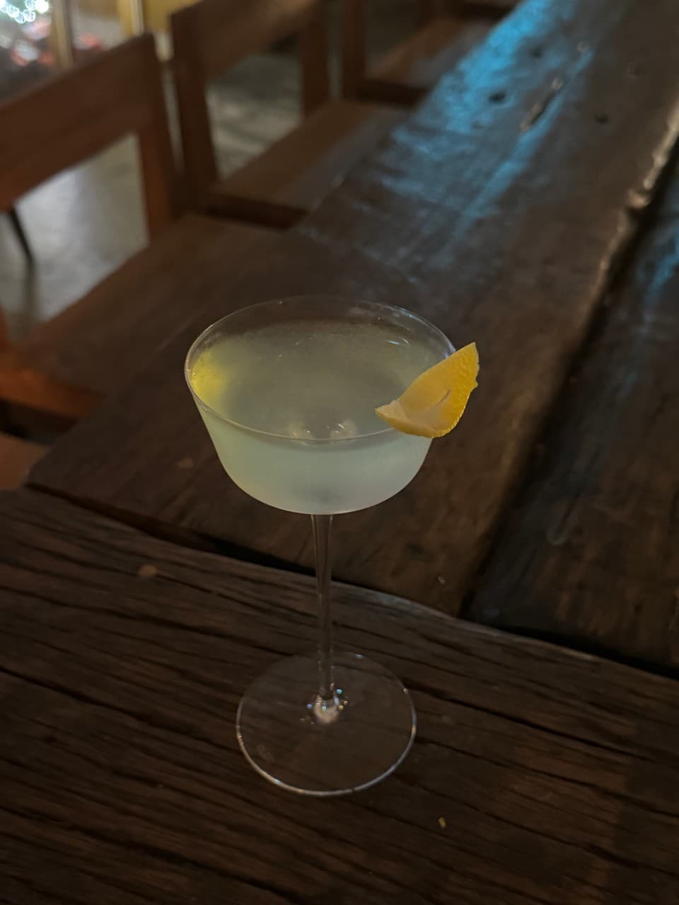 어두운 실내 나무 카운터 석에 놓인 레몬 껍질이 장식된 투명한 칵테일 잔과 비어있는 1인용 좌석