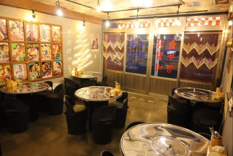 황색 전구 조명 아래 원형 스테인리스 테이블과 검은색 원통형 의자가 배치된 내부. 벽면의 고전 영화 포스터와 트리 형태의 전구 장식