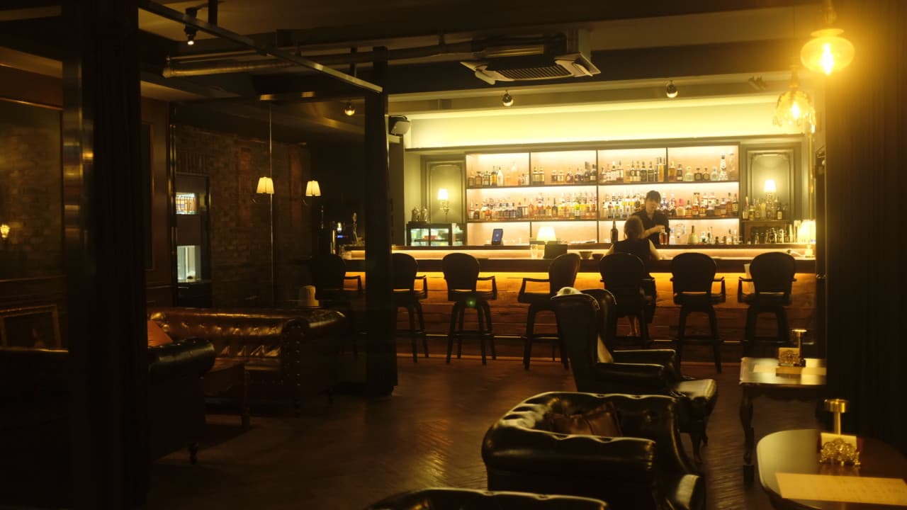 노란 조명이 비치는 술병 진열장과 긴 나무 바 테이블에 일렬로 배치된 1인용 높은 의자 및 가죽 소파 전경