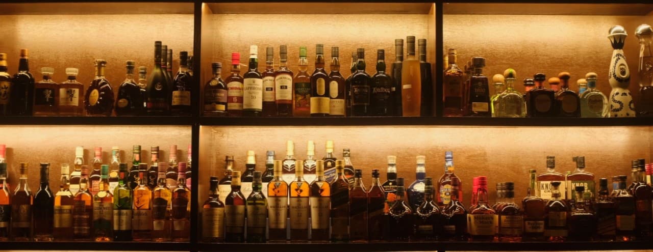 황금빛 간접 조명이 들어오는 2단 진열 선반 위에 일렬로 빽빽하게 놓인 다양한 위스키와 양주 병들