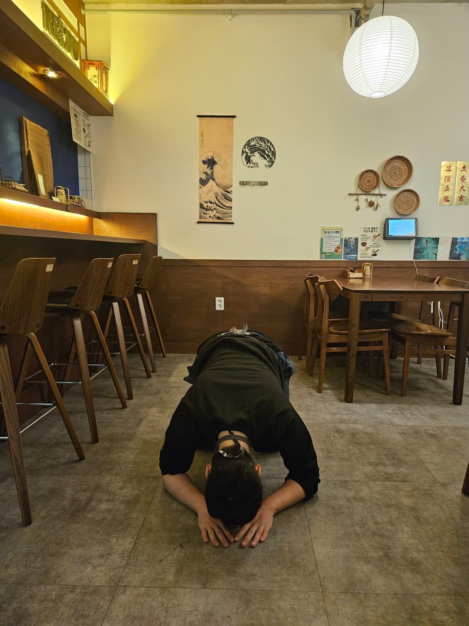목재 카운터석과 1인용 의자가 배치된 술집 내부 바닥에서 큰절을 하는 인물과 벽면의 일식 족자