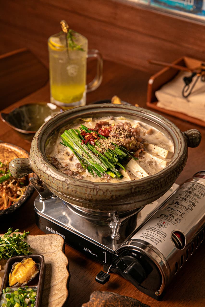 목재 테이블 위 휴대용 버너에 놓인 고기와 두부, 부추가 담긴 전골 냄비와 레몬 조각을 띄운 하이볼 한 잔