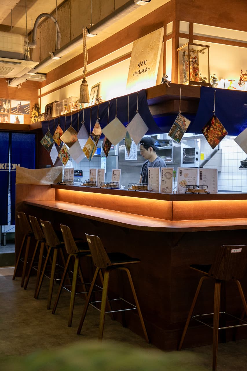 주황색 간접 조명이 들어오는 긴 목재 카운터 테이블과 높은 바 의자가 배치된 1인 중심의 좌석 공간